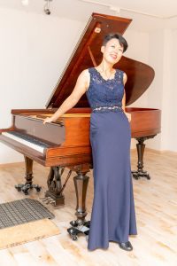 Woman in blue dress standing in front of a brown piano.