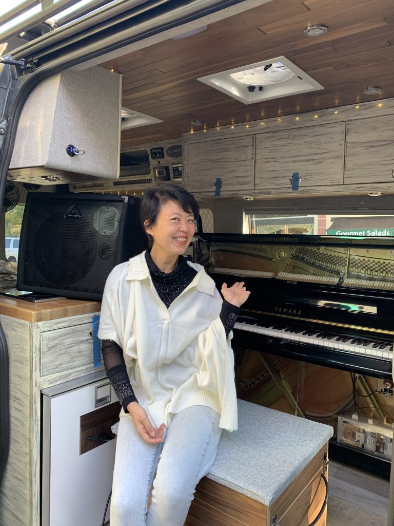 woman sitting at the entrance of a van smiling with a piano (in the van) behind her