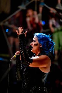 Woman with blue hair black leather arm bands and corset singing in a microphone, blurry human faces in background