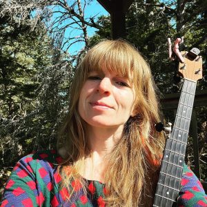 Strawberry blond woman facing camera holding a banjo in front of trees.