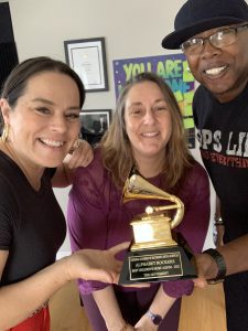 3 Smiling people huddling around the Alphabet Rockers' 2023 Grammy award statue