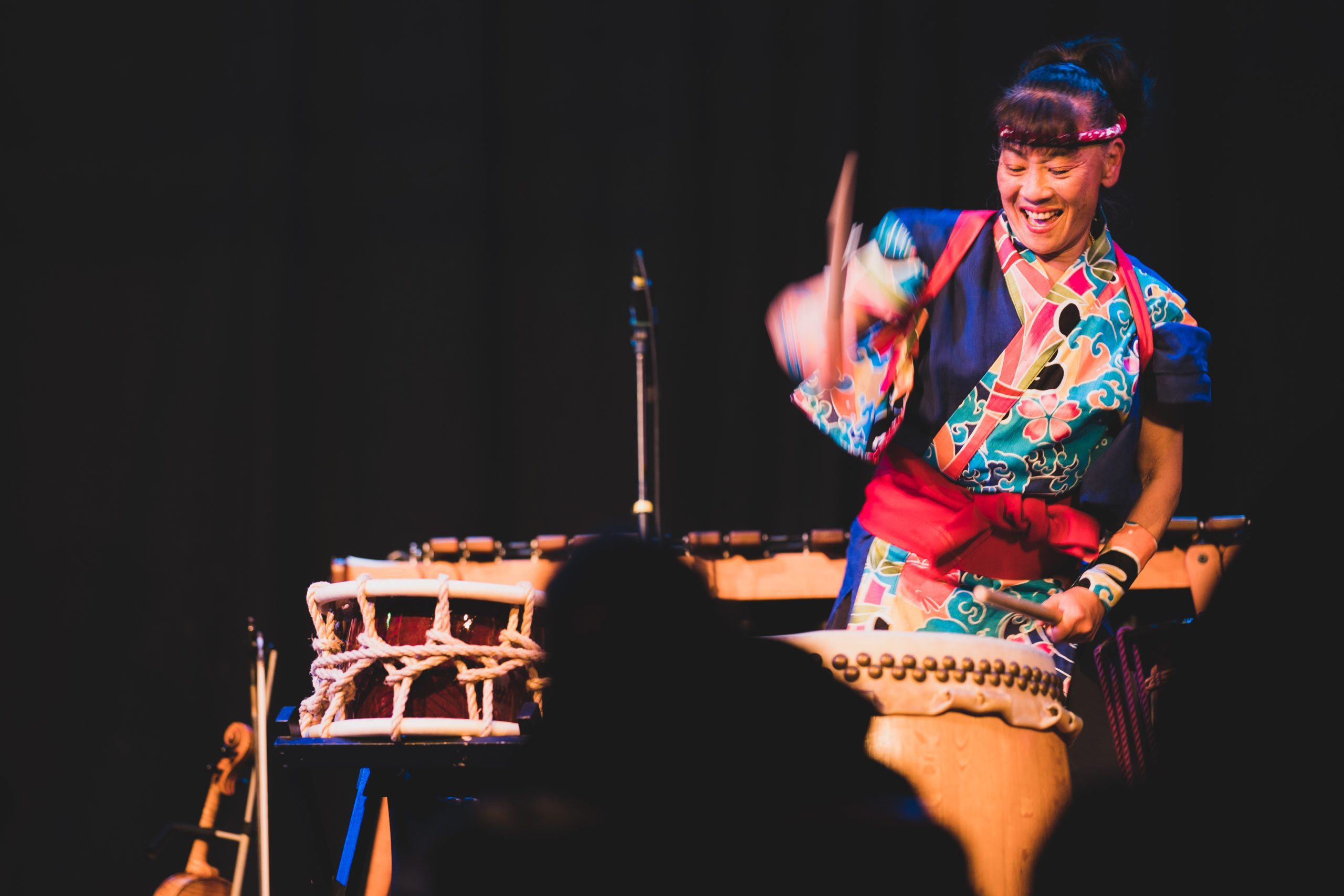woman in colorful outfit playing a big drum with bachi (hardwood drum sticks)