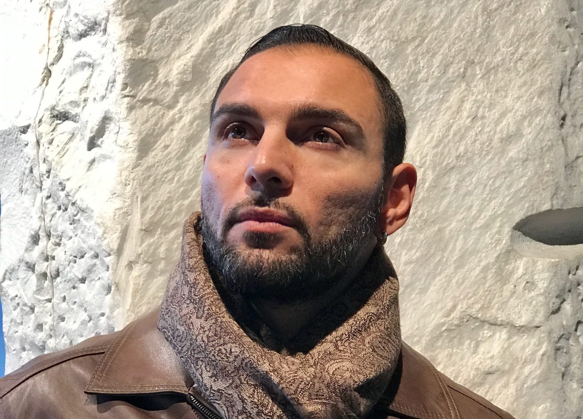 Dark haired and eye man looking slightly to the side of camera wearing brown scarf and brown leather jacket.