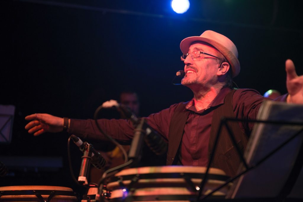 Man in hat with face microphone in front of drums with his arms out wearing glasses