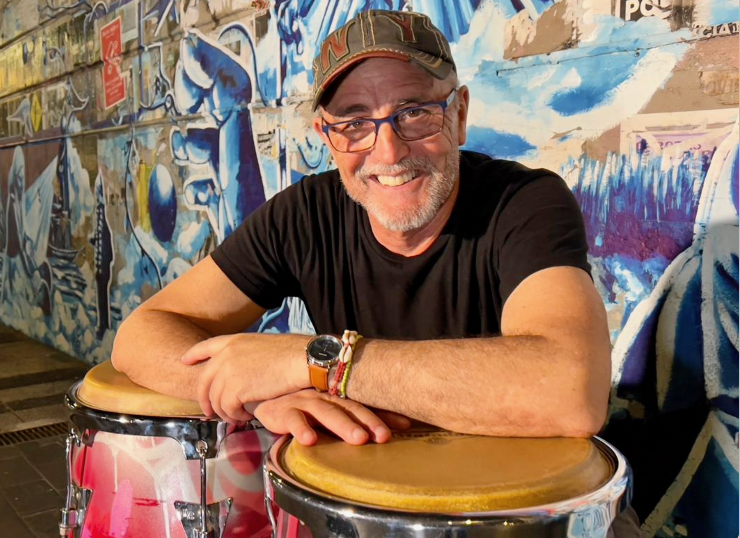 Man smiling and leaning on conga drums. Man is wearing a hat and glasses