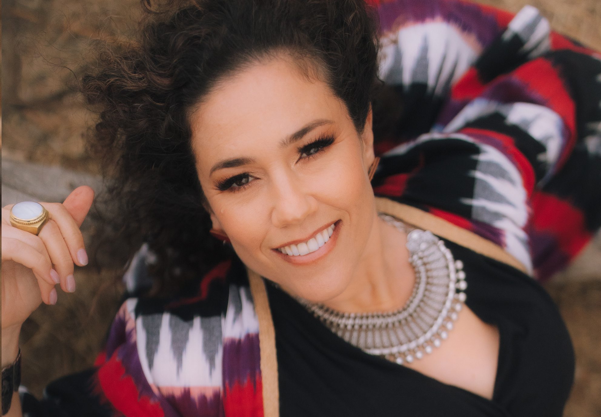 Brown curly-haired woman looking up at camera wearing silver necklace, black dress and red, white and black shawl