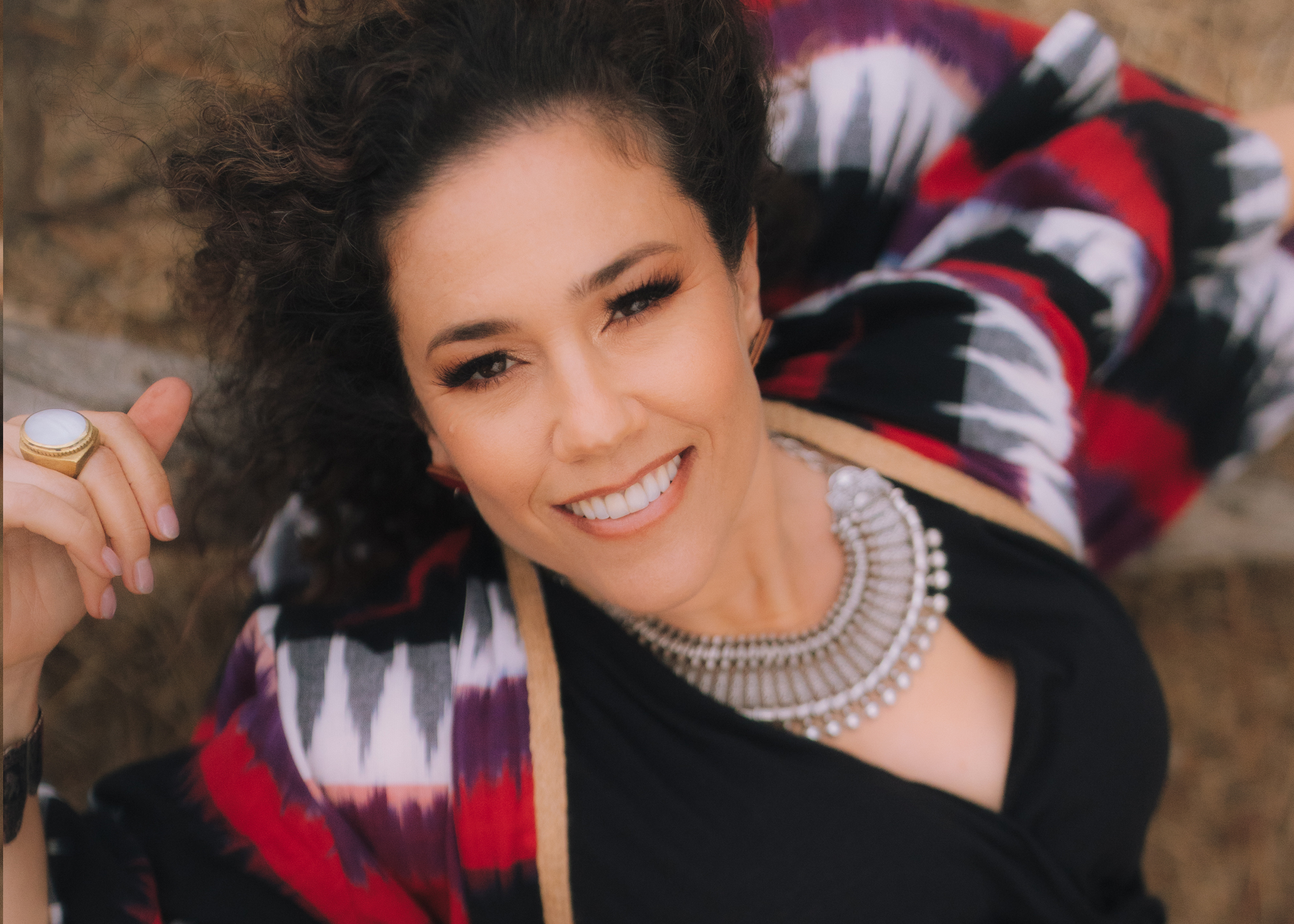 Brown curly-haired woman looking up at camera wearing silver necklace, black dress and red, white and black shawl