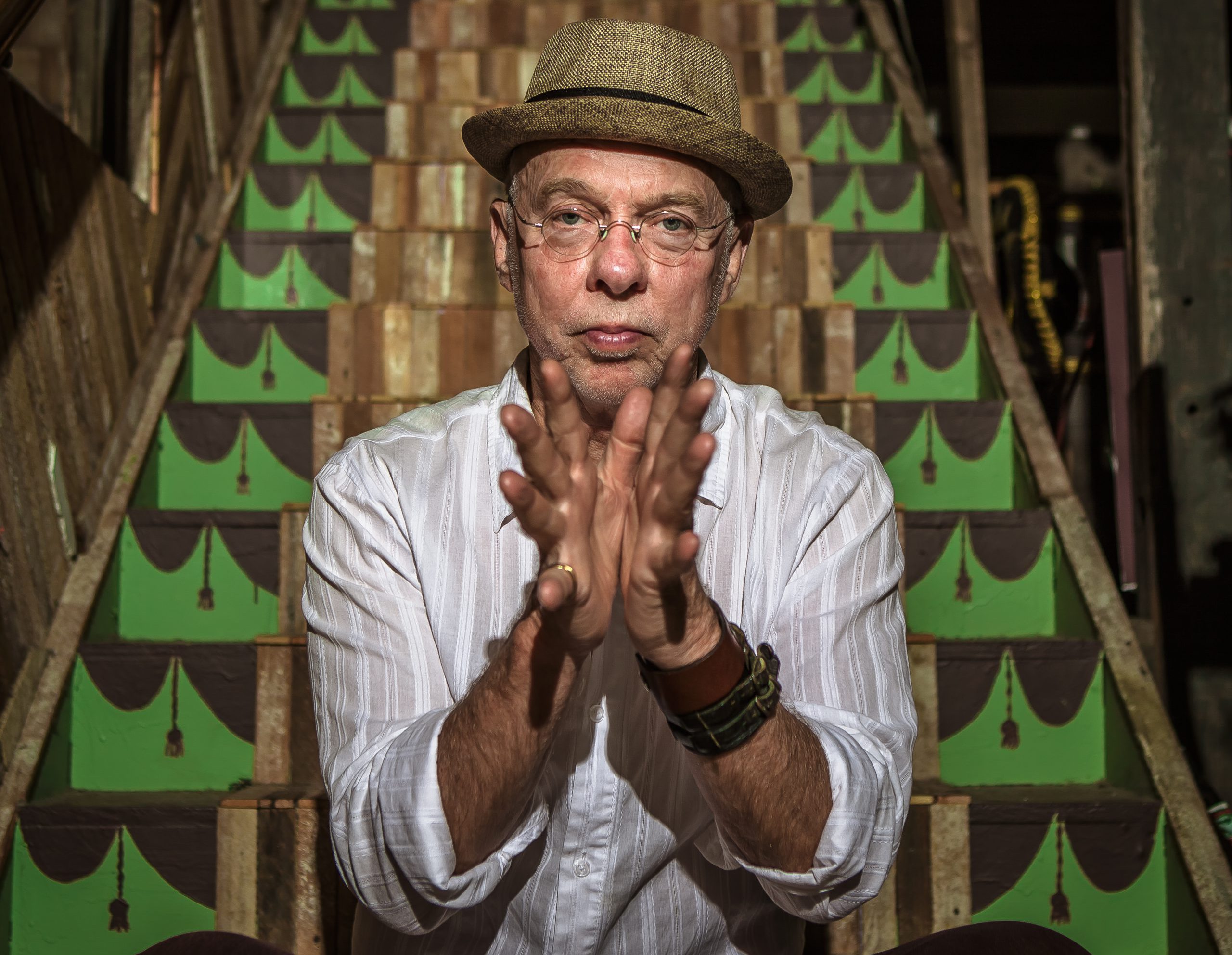 Man in hat and white shirt clapping hands together seated on stairs with a green pattern.