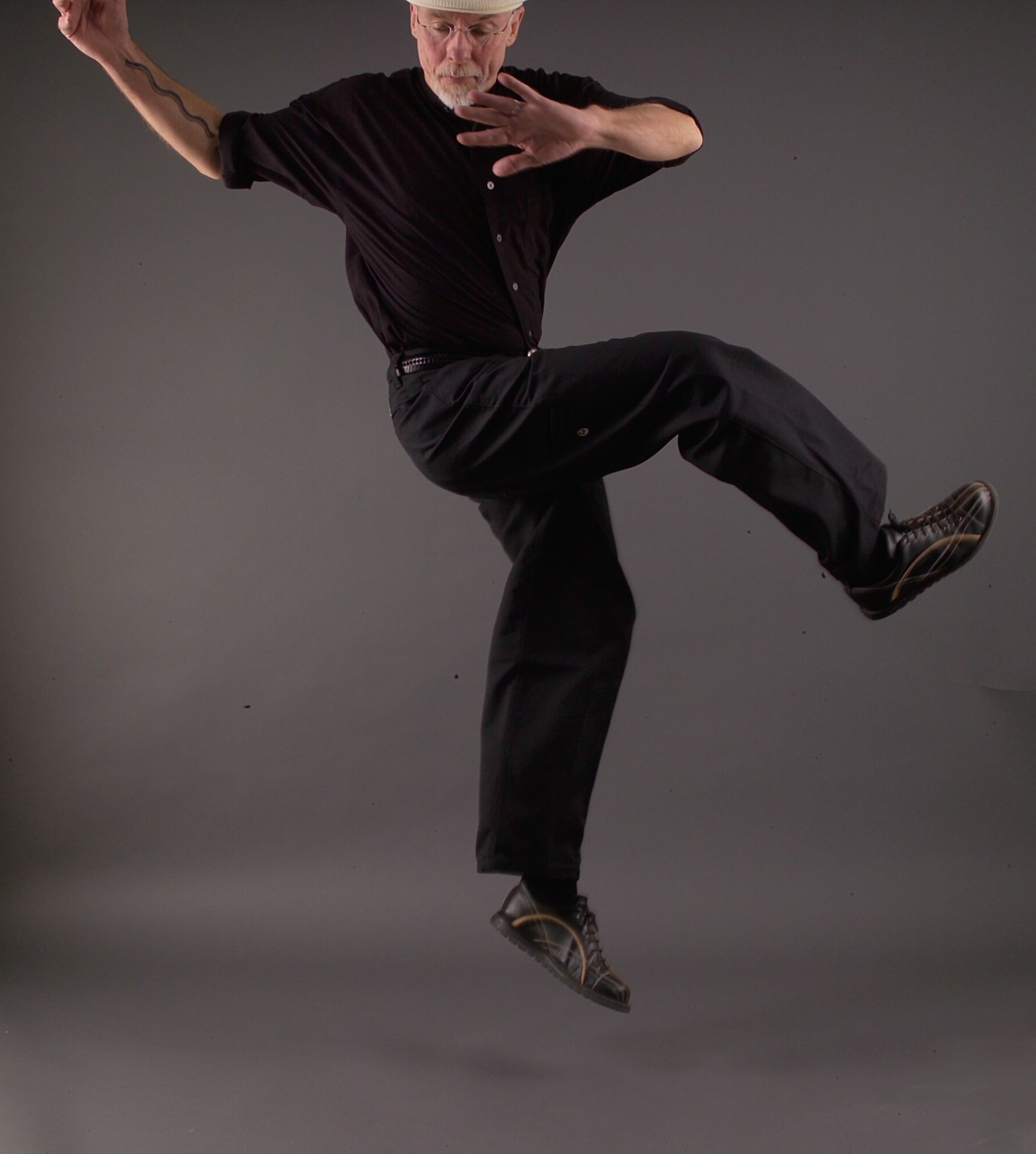 Keith Terry in a black pants suit and shirt leaping off the ground with his arms up and looking behind himself.